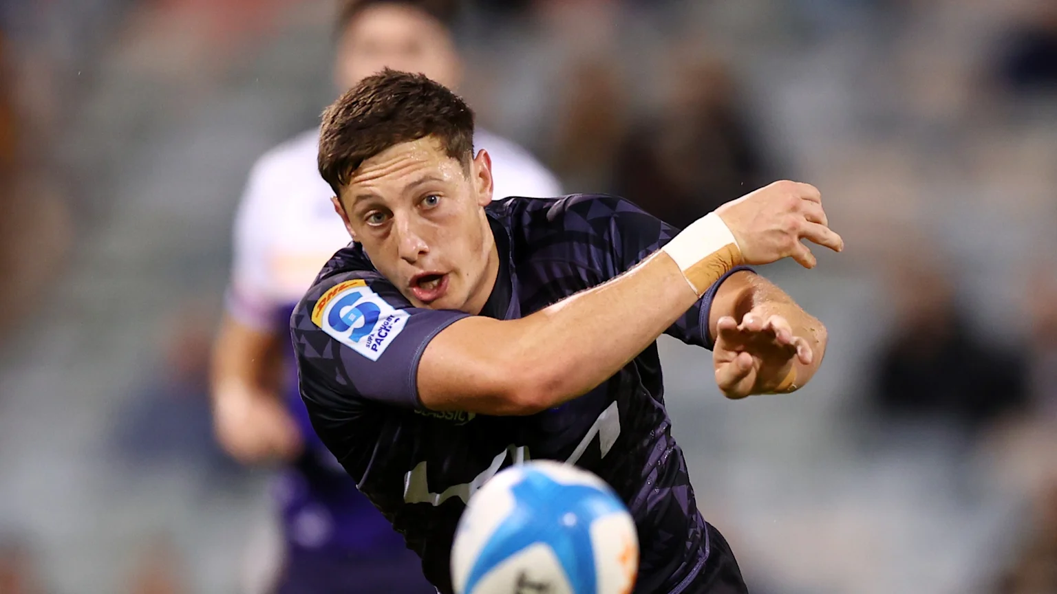 A young male rugby player in a dark jersey with a Super Rugby Pacific logo on his shoulder looks intently while in action, with a rugby ball in the foreground.
