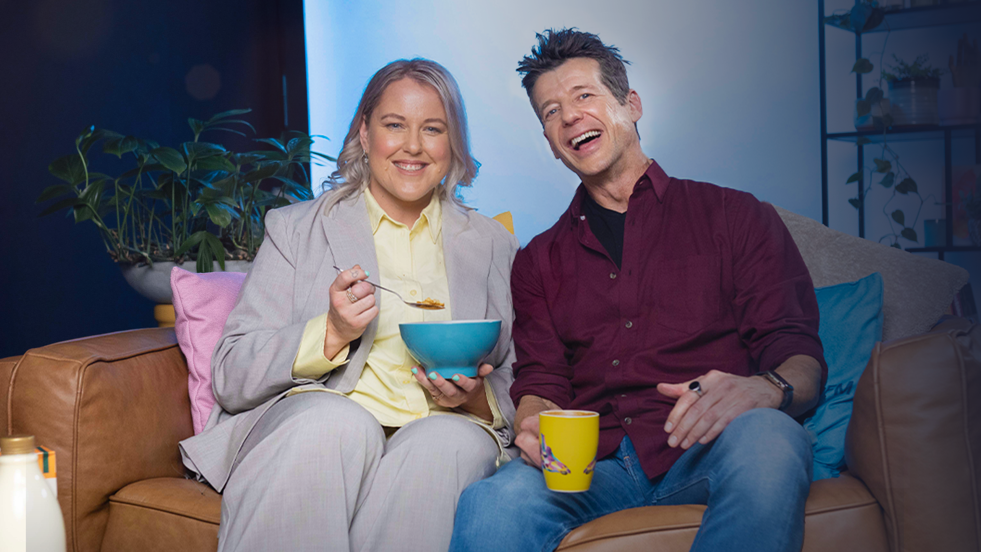 Lana and Simon sitting on a couch, smiling at the camera. Lana is holding a bowl of food, and Simon has a yellow mug in his hand.