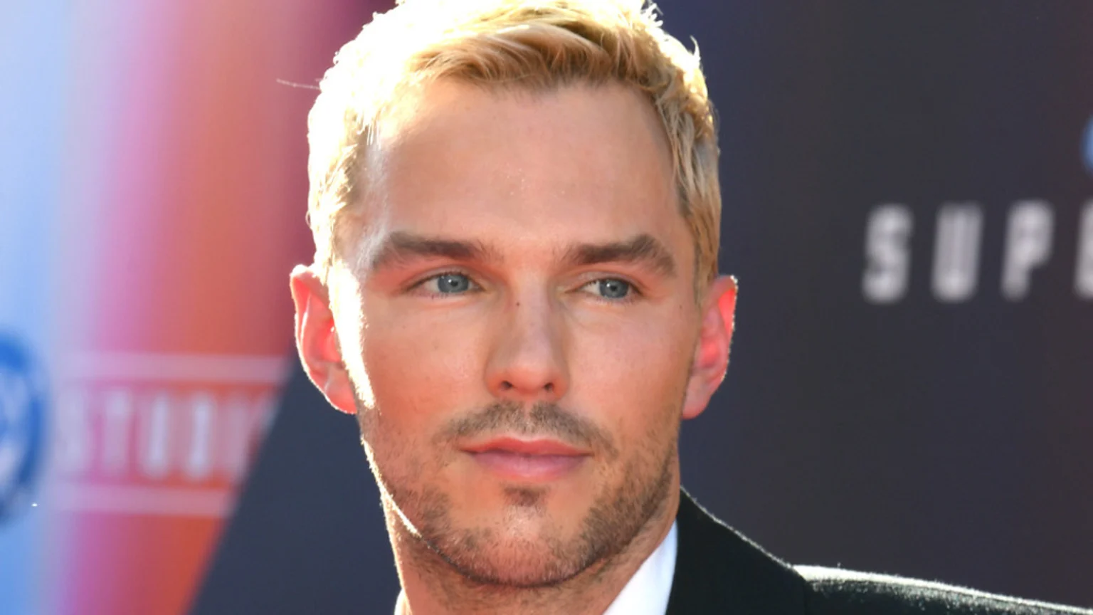 A close-up shows actor Nicholas Hoult with blonde hair, blue eyes, and light stubble, looking left with a subtle smile, against a blurred background with soft colors and "SUPER" text.