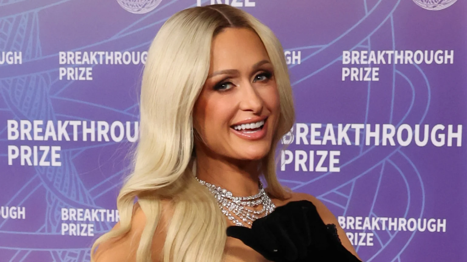 Paris Hilton with long blonde hair, smiling at the camera, wearing a diamond necklace and a black glove, against a purple background with "BREAKTHROUGH PRIZE" text.