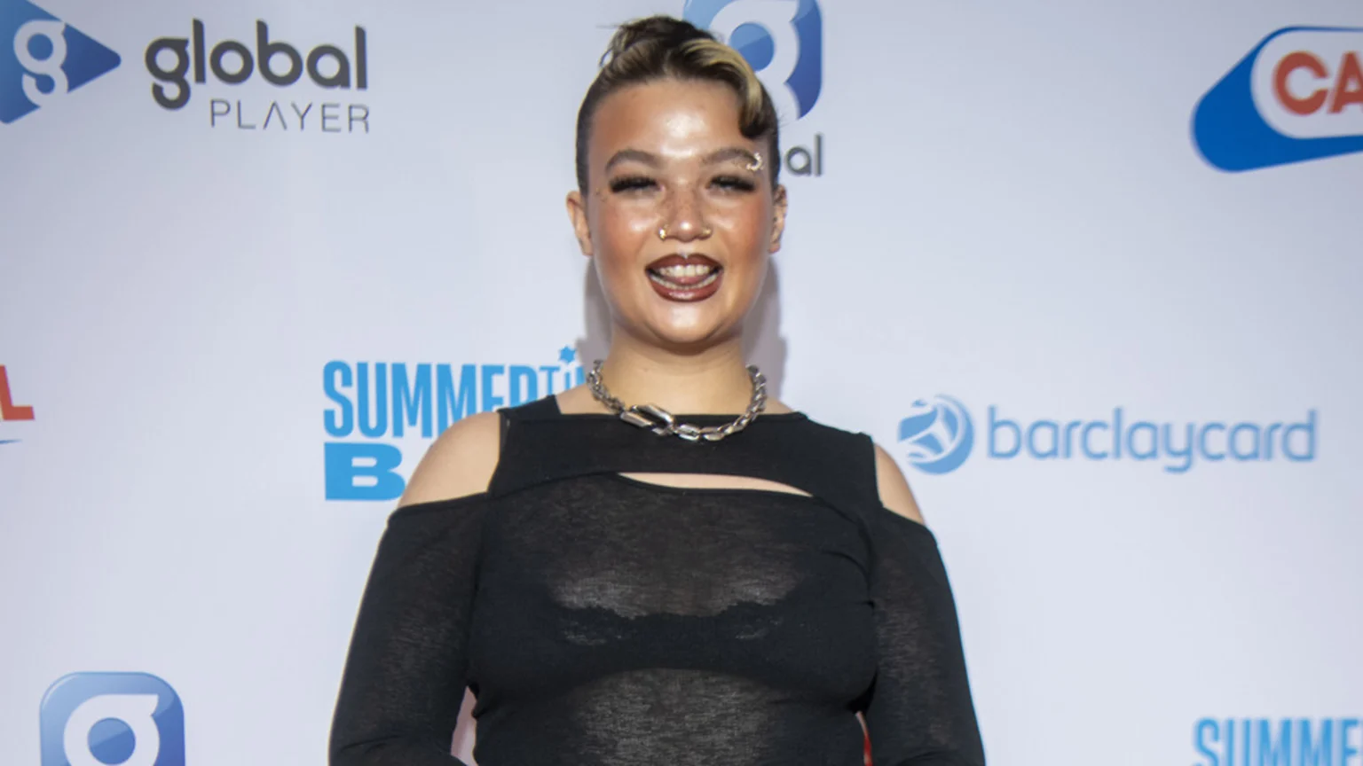 Lola Young, a woman with dark hair, eyebrow and nose piercings, and dark lipstick, smiles at the camera, wearing a black top and silver chain necklace against a white backdrop with logos.