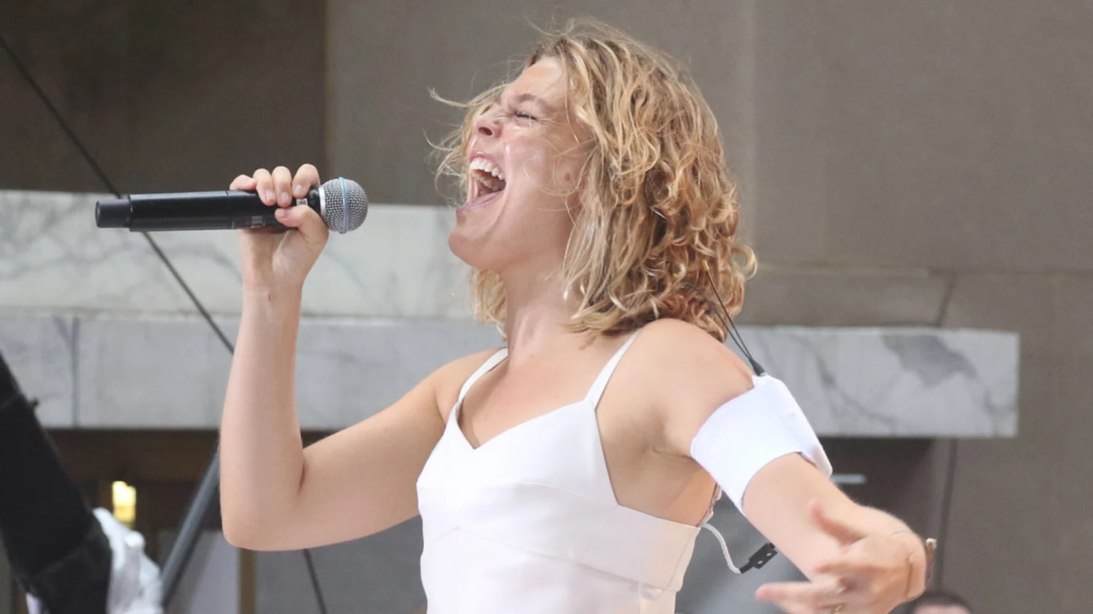 A woman with curly blonde hair in a white top sings passionately into a microphone with eyes closed and mouth wide open, an armband with wires visible on her left arm.
