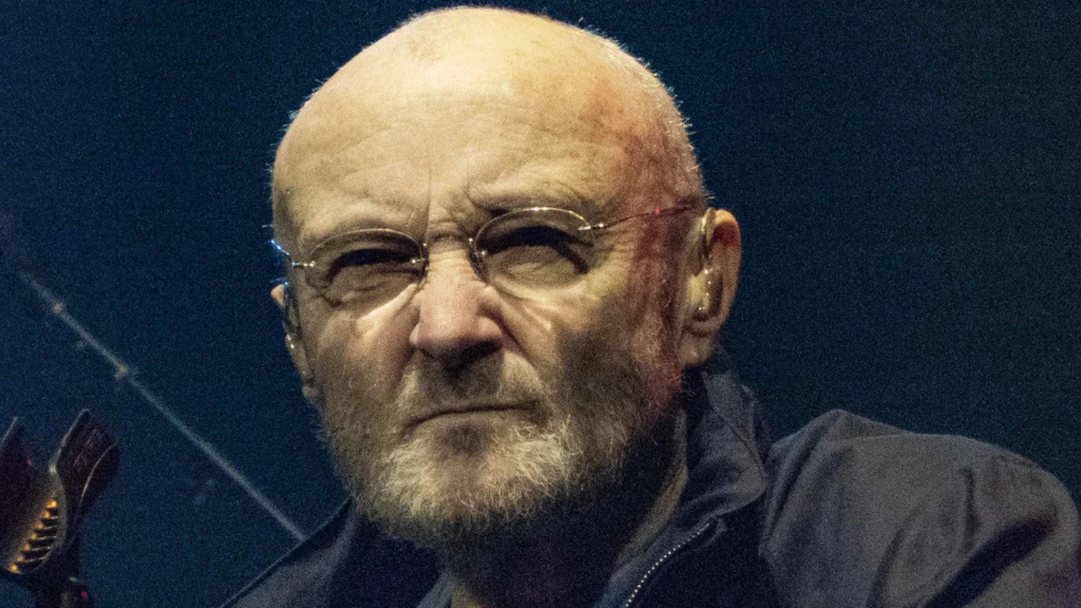 A close-up of Phil Collins, a bald man with glasses and a gray beard, looking intently at the camera with a somber expression in a dimly lit setting, wearing a dark jacket.