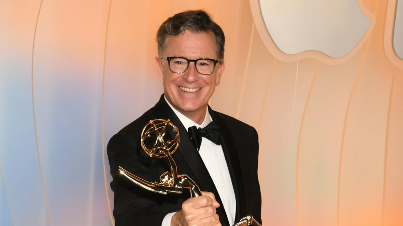 Stephen Colbert, in a black tuxedo and glasses, smiles brightly as he holds up a golden Emmy award against a backdrop of orange and blue tones with a partial white Apple logo.