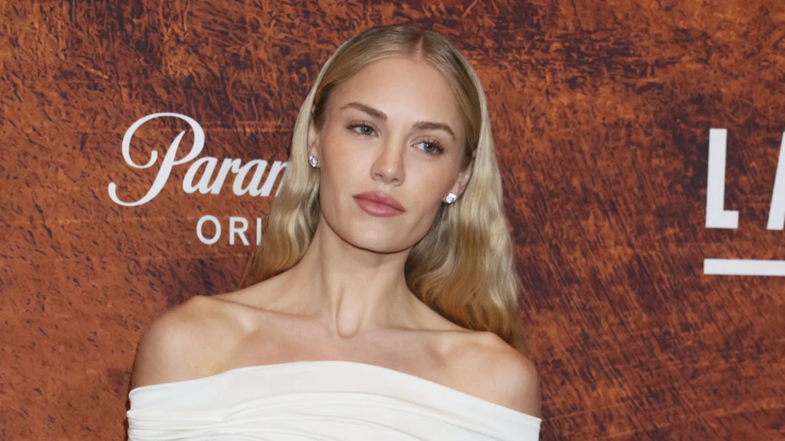Michelle Randolph, with long blonde hair and stud earrings, wears an off-the-shoulder white top, looking slightly left against a brown background with "Paramount" and "LANDMAN" logos.