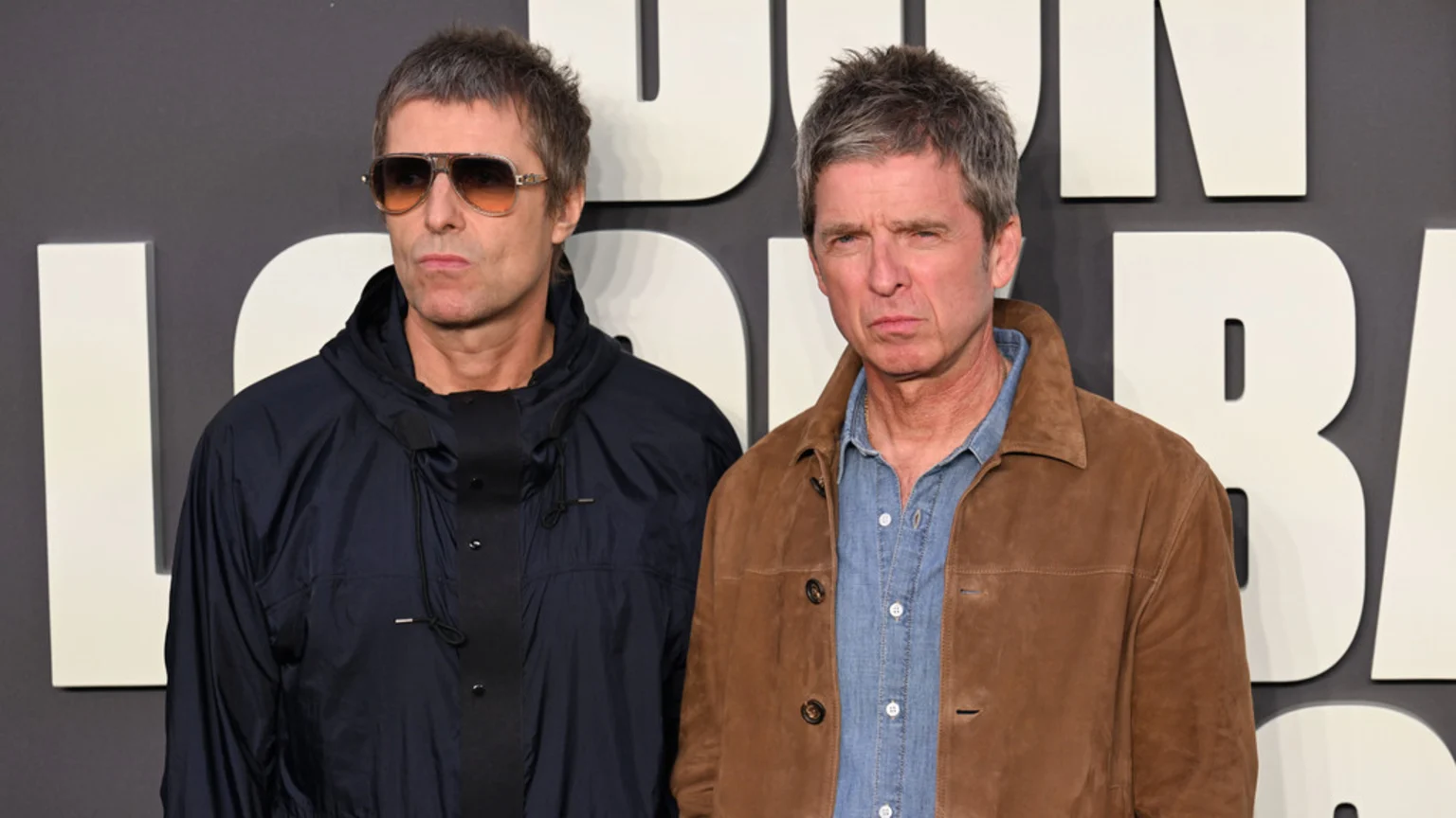Liam Gallagher in sunglasses and a dark jacket stands beside Noel Gallagher in a brown jacket and denim shirt, both with serious expressions, against a background of large white letters.