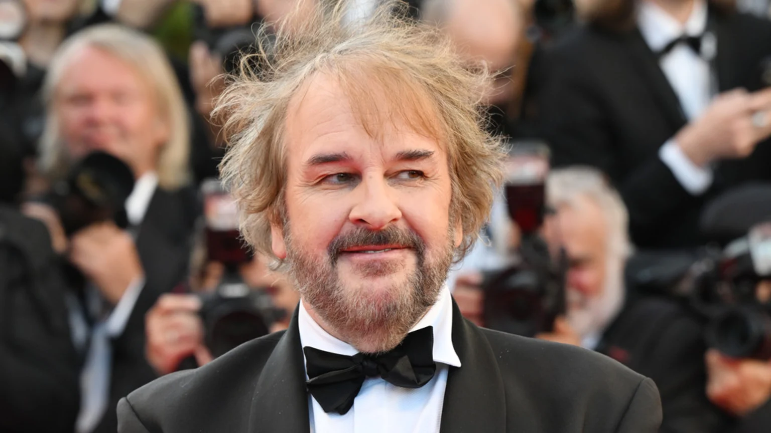 A man identified as Peter Jackson with messy light brown hair and a beard, wearing a black tuxedo and bow tie, smiles slightly while looking to his right.