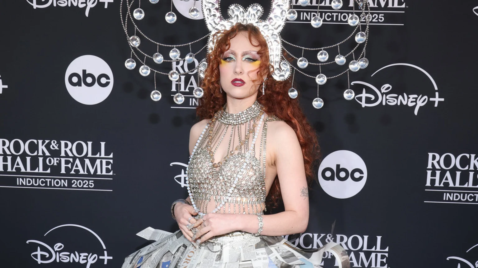 A person with long red hair, a sparkling crown, and dramatic makeup wears an elaborate, beaded silver top and a newspaper skirt on a black background with "Rock & Roll Hall of Fame" logos.