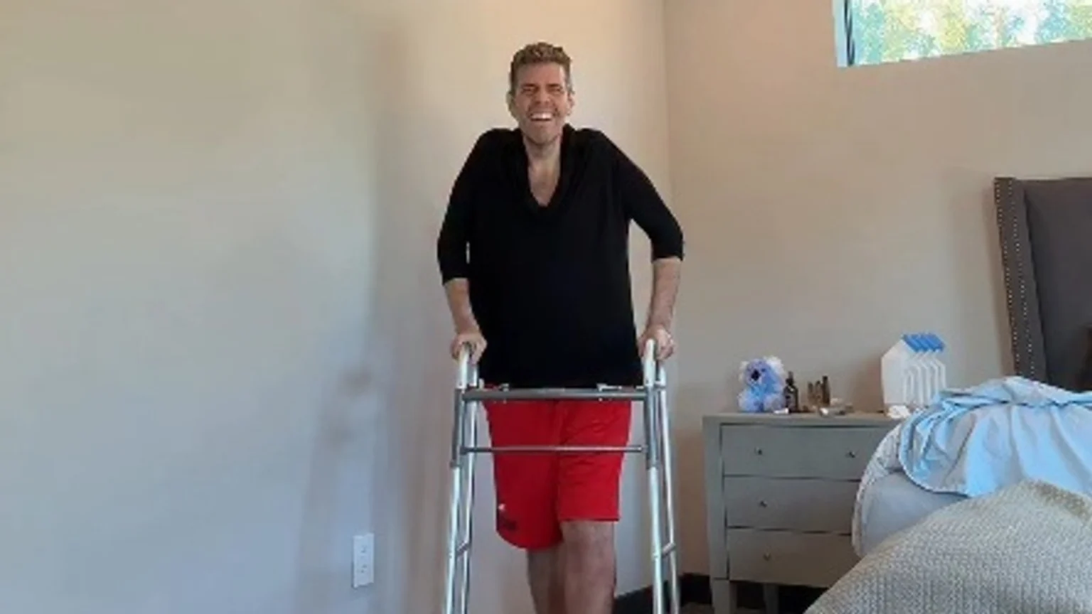 Perez Hilton stands, smiling broadly, holding a walker as he walks again, dressed in a black top and red shorts, in a room.