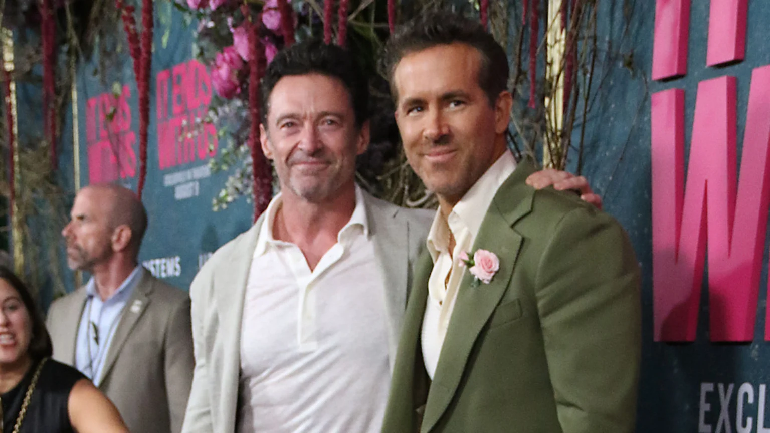 Hugh Jackman and Ryan Reynolds smile at the camera, with Reynolds' arm around Jackman's shoulder, in front of a backdrop reading "DEADPOOL & WOLVERINE."