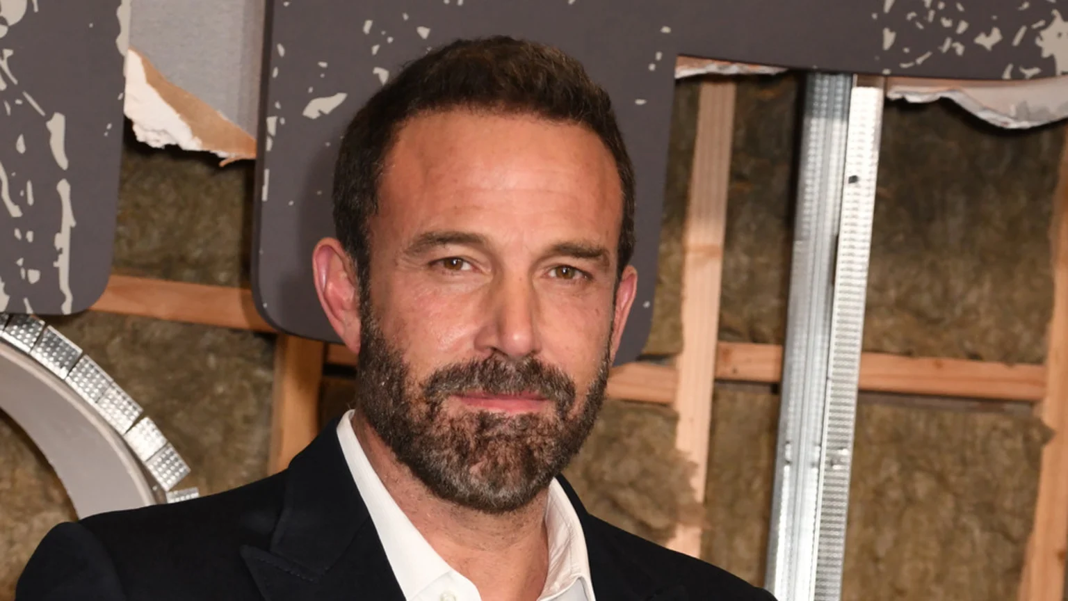 Ben Affleck, with a beard and short dark hair, looks at the camera wearing a dark suit against a background of exposed wall framing, insulation, and broken grey panels.