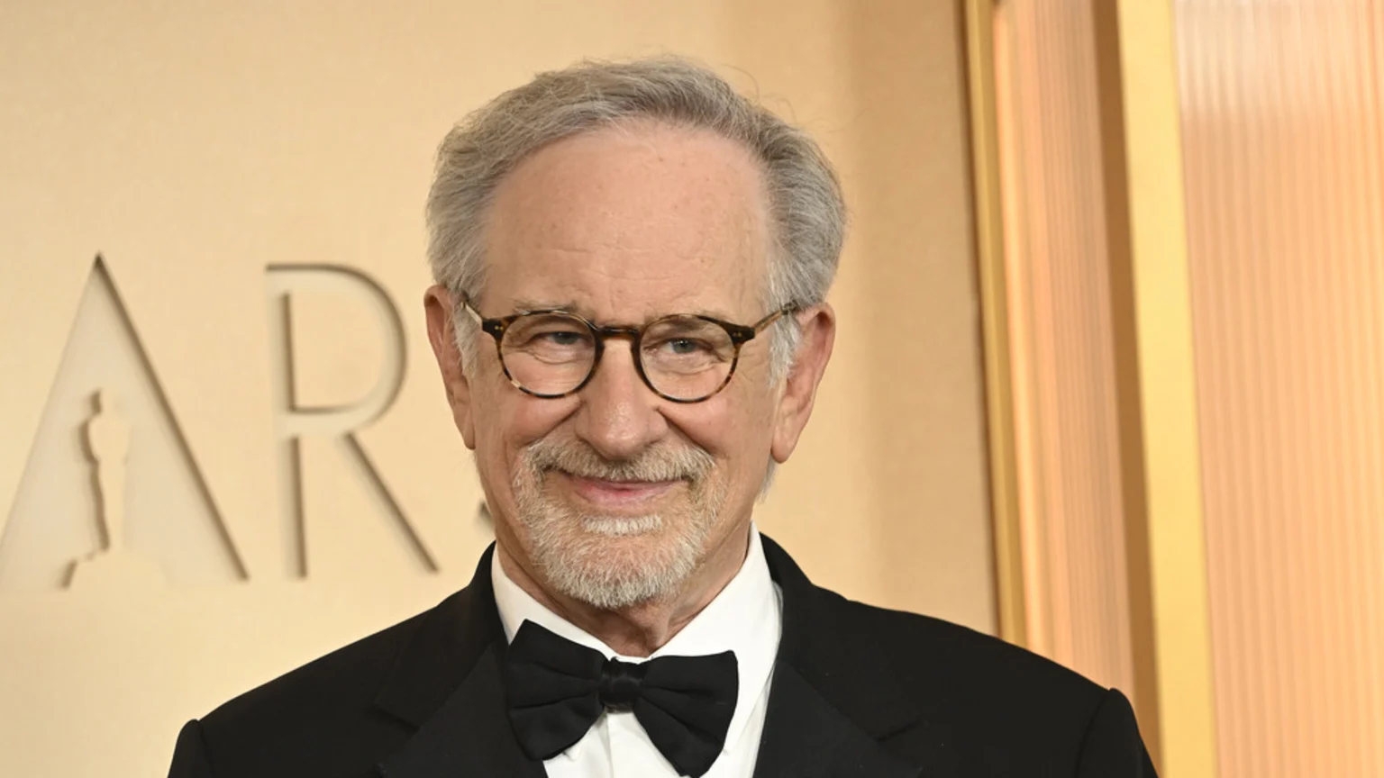 Steven Spielberg smiles, wearing glasses, a black tuxedo, and a bow tie, against a gold background showing part of the word "ARS" with an Oscar silhouette.