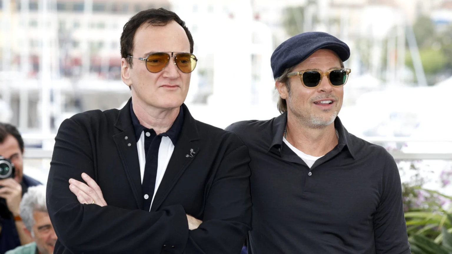 Quentin Tarantino in a blazer and yellow-tinted sunglasses stands next to Brad Pitt in a blue cap, sunglasses, and black polo shirt, both posing outdoors.