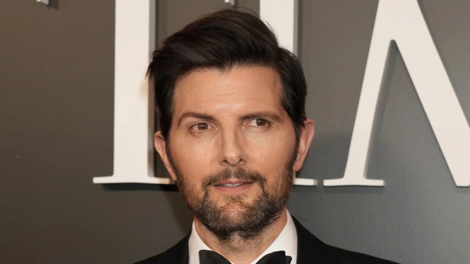 Actor Adam Scott, a man with dark hair and a beard, wears a tuxedo with a bow tie and a subtle smile, looking slightly right against a dark grey wall with white letters.