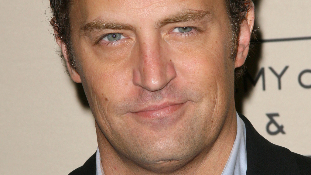 Close-up portrait of actor Matthew Perry smiling subtly, looking at the camera. He wears a light blue shirt and dark jacket against a light, blurred background.