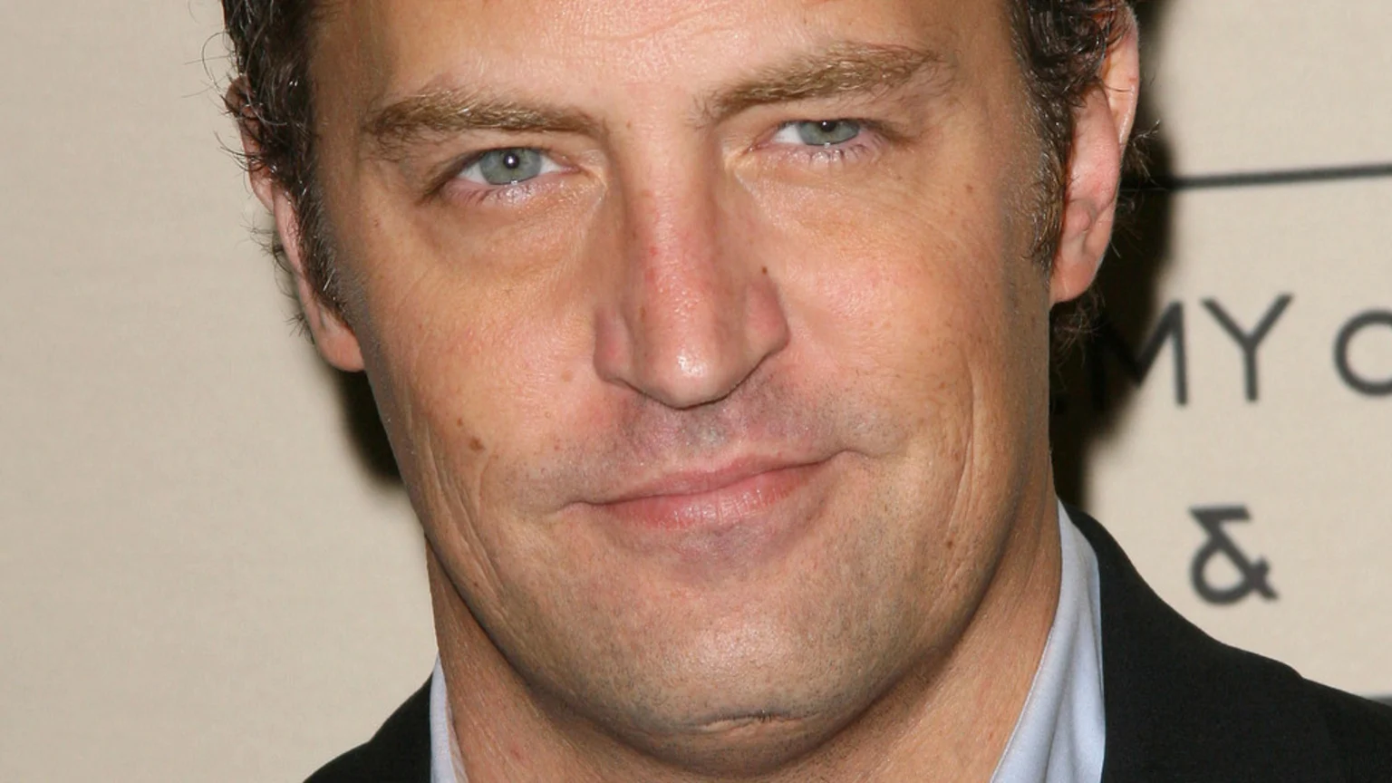 Close-up portrait of actor Matthew Perry smiling subtly, looking at the camera. He wears a light blue shirt and dark jacket against a light, blurred background.