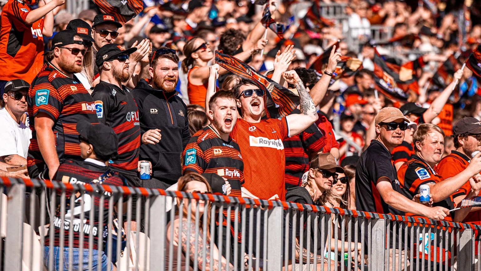 A large crowd of excited fans, many in black and red team colors, cheer and wave flags from the stands of a sunny outdoor stadium.