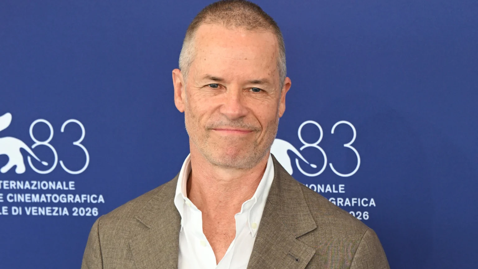 A man with short grey hair and a light beard smiles in a brown suit jacket and white shirt against a blue backdrop with the white logo and text for the 83rd International Film Festival of Venice 2026.