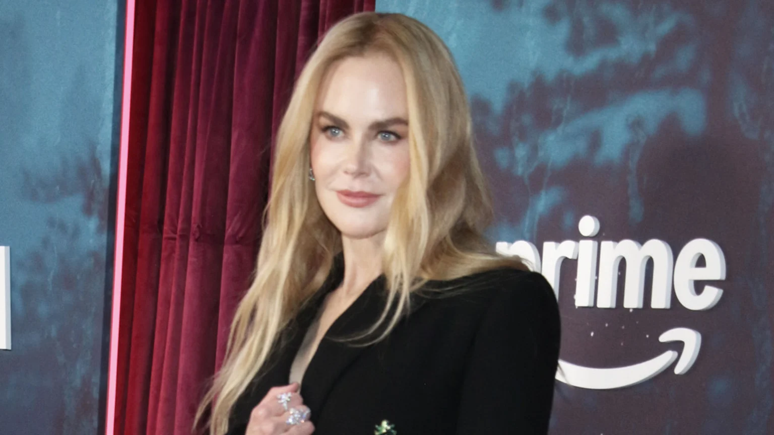 Actress Nicole Kidman with long blonde hair, wearing a black blazer and rings, stands against a background with red curtains and an Amazon Prime logo.
