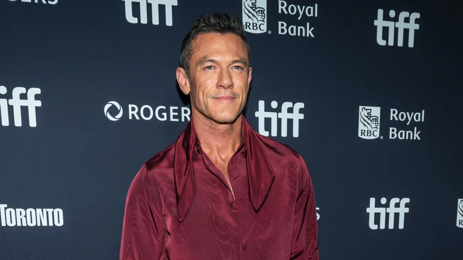 Luke Evans, a man with short dark hair and a dark red satin shirt, smiles slightly in front of a dark backdrop with white "tiff", "RBC Royal Bank", and "ROGERS" logos.