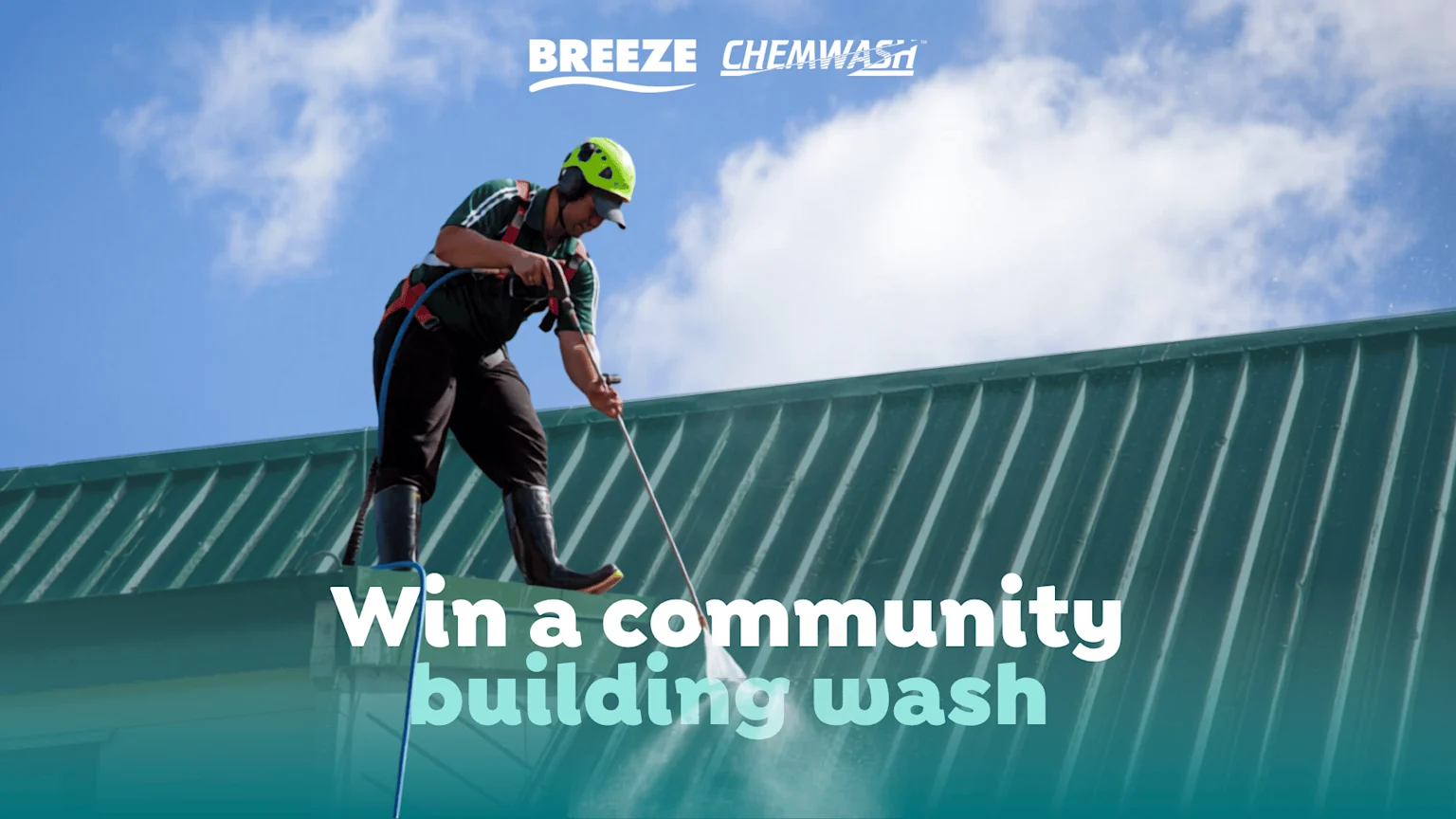 A person in safety gear pressure washing a green corrugated roof under a blue sky, with "BREEZE CHEMWASH" logos and text "Win a community building wash".