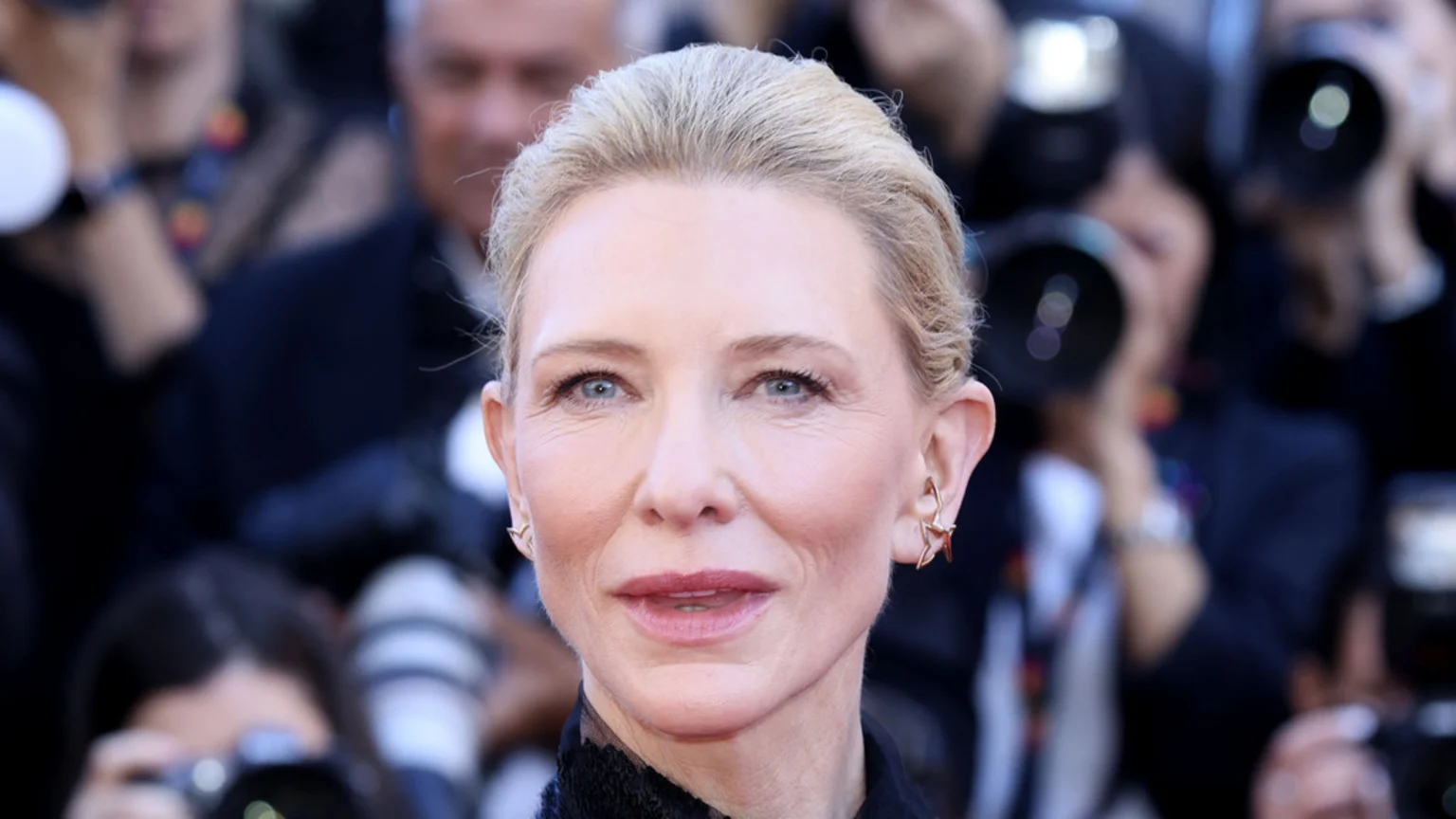 Cate Blanchett smiles with her blonde hair pulled back, wearing star-shaped ear cuffs, while blurred photographers are visible in the background.