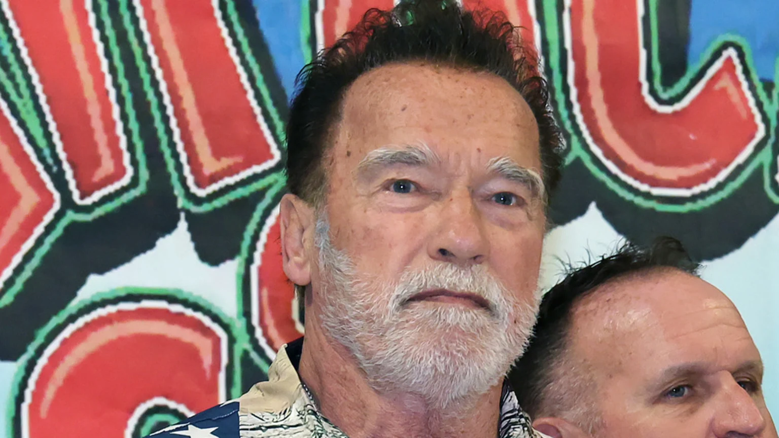 Close-up of Arnold Schwarzenegger with a gray beard and dark hair, wearing a patterned shirt, looking directly, with a colorful background and another person partially visible to the lower right.