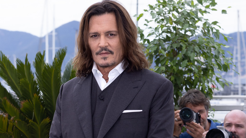 Johnny Depp, a man with long brown hair and a mustache, wears a dark suit and white shirt, looking at the camera with green plants and a marina in the background.