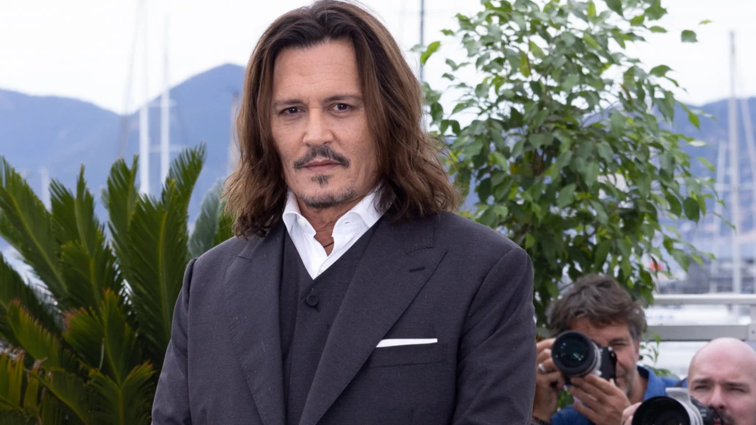 Johnny Depp, a man with long brown hair and a mustache, wears a dark suit and white shirt, looking at the camera with green plants and a marina in the background.