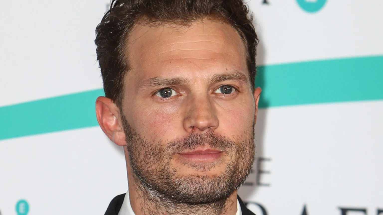 Close-up of actor Jamie Dornan, with dark wavy hair and a short, scruffy beard, looking forward with blue eyes against a white and turquoise background.