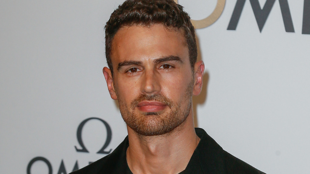 Theo James, a man with short brown curly hair and a beard, looks directly forward, wearing a dark shirt against a white background with an Omega logo.