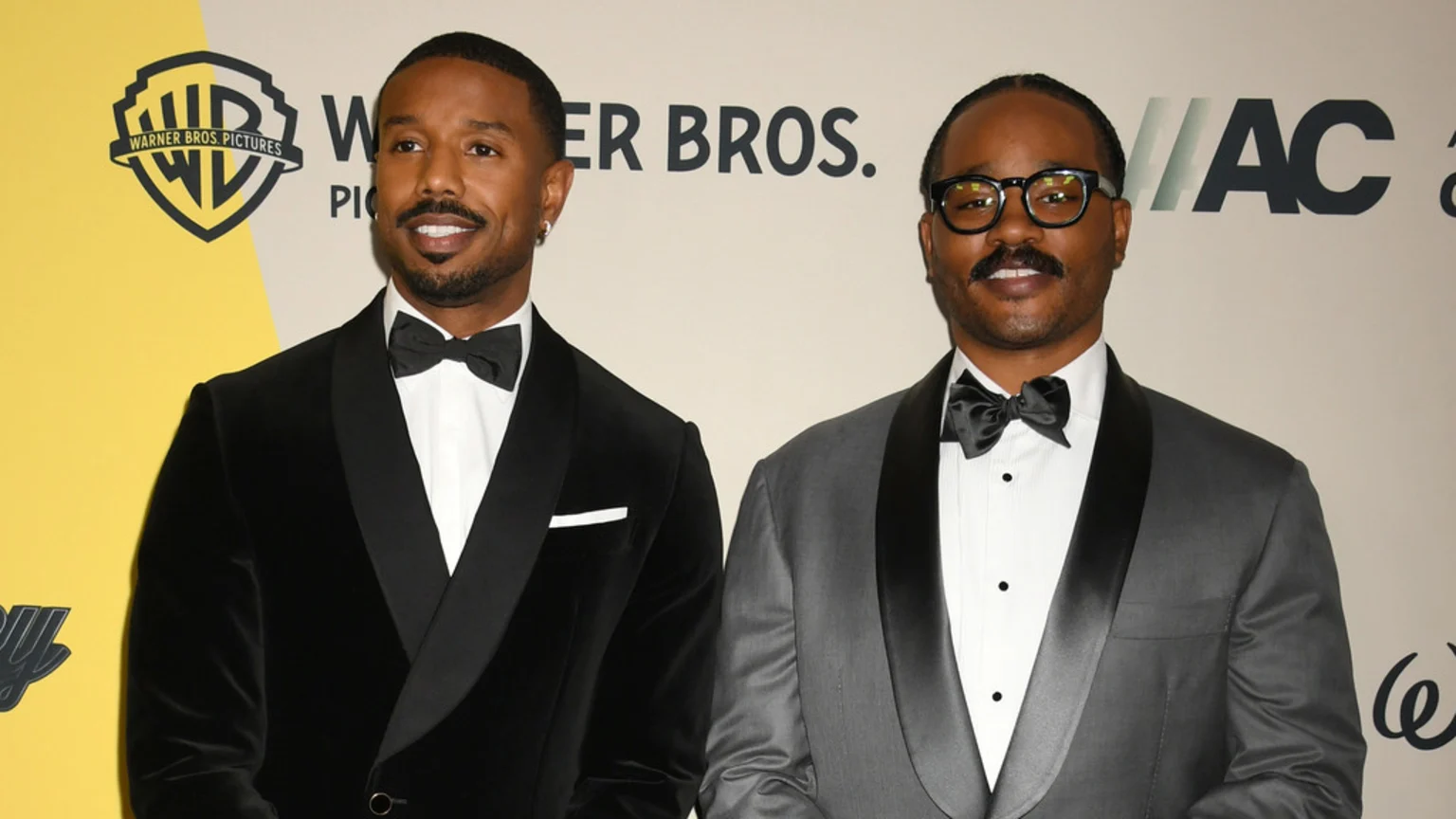 Michael B. Jordan in a black velvet tuxedo and Ryan Coogler in a grey tuxedo with glasses smile while standing side-by-side in front of a Warner Bros. logo.
