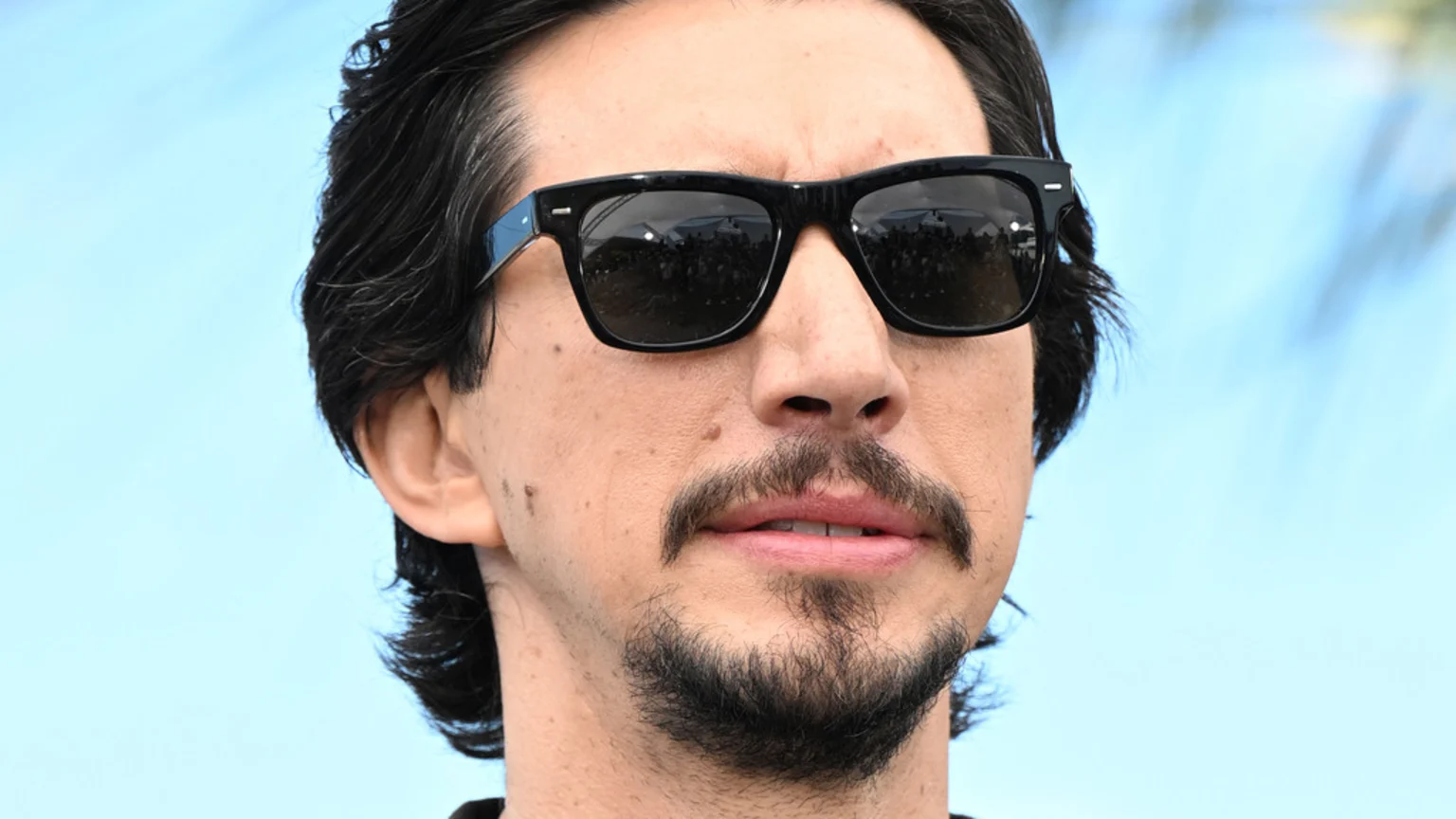 Actor Adam Driver with dark hair, a goatee, and mustache, wears black sunglasses reflecting a crowd, against a bright blue sky.