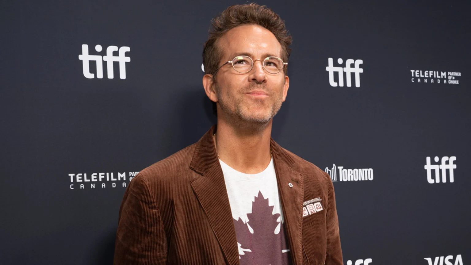 Ryan Reynolds, wearing glasses, a brown corduroy jacket, and a maple leaf t-shirt, looks up against a dark background with white "tiff" and "TELEFILM CANADA" logos.