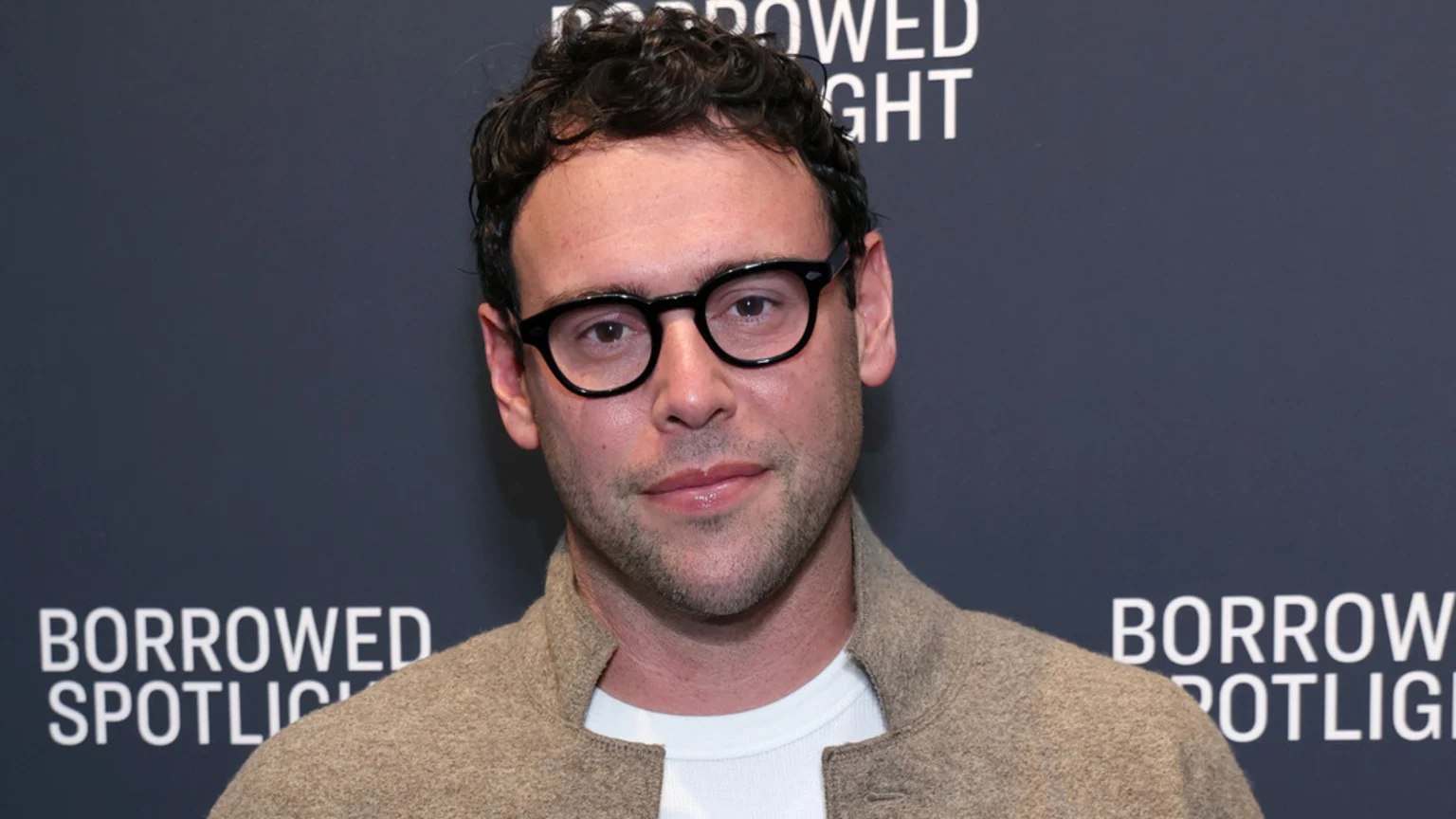 Scooter Braun, a man with dark curly hair, black round glasses, and light facial hair, wears a tan jacket over a white shirt, smiling slightly against a dark background.