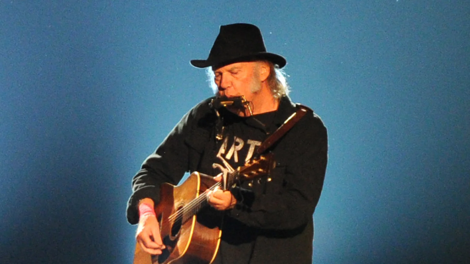 Musician Neil Young, wearing a black hat, black shirt with "ART" visible, and a pink wristband, plays an acoustic guitar while simultaneously playing a harmonica on a neck rack against a blue background.