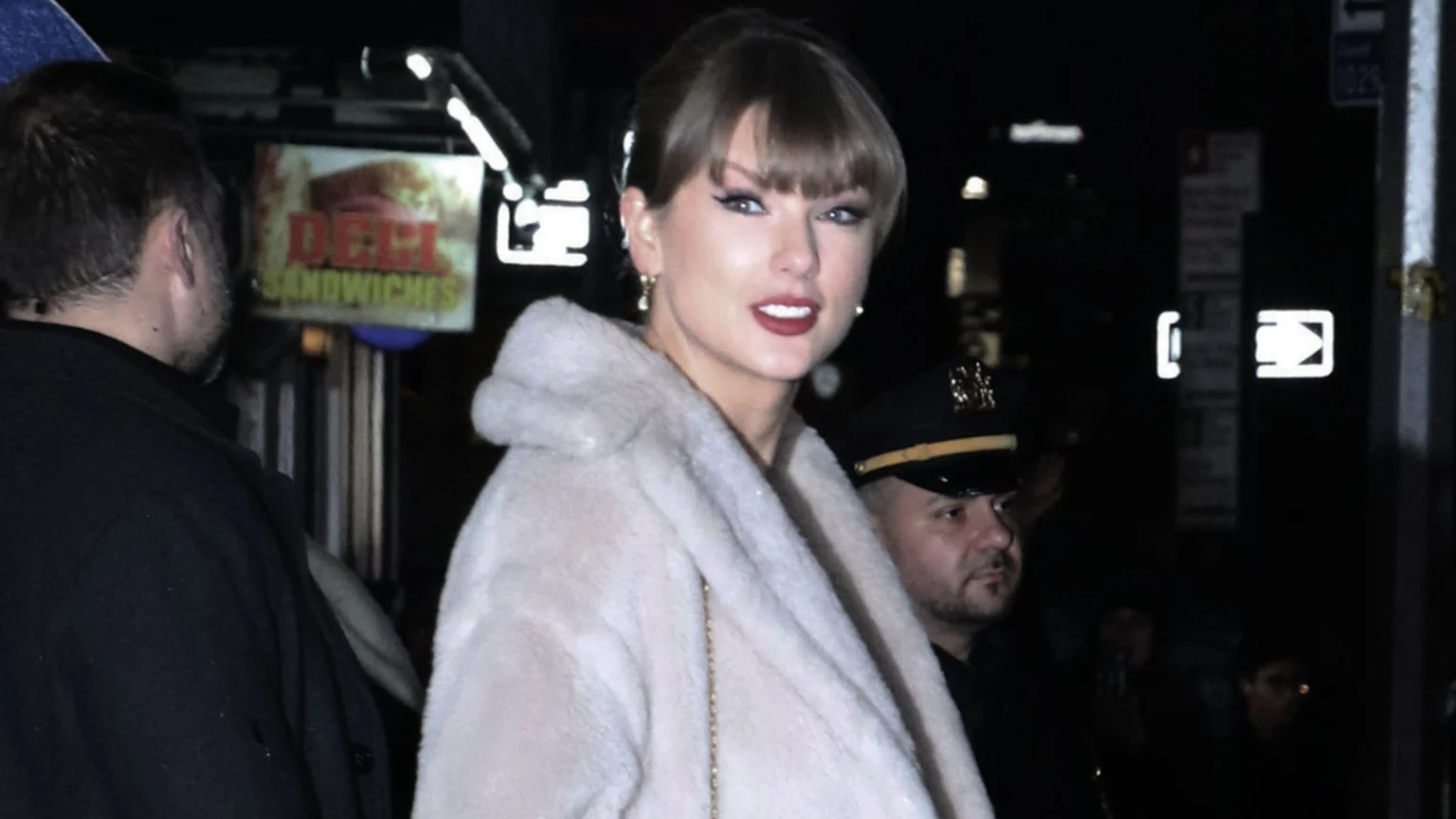 Taylor Swift smiles, wearing a light faux fur coat, with red lipstick and bangs, looking towards the viewer. A man in a dark uniform is partially visible next to her, and a "DELI SANDWICHES" sign is in the background.