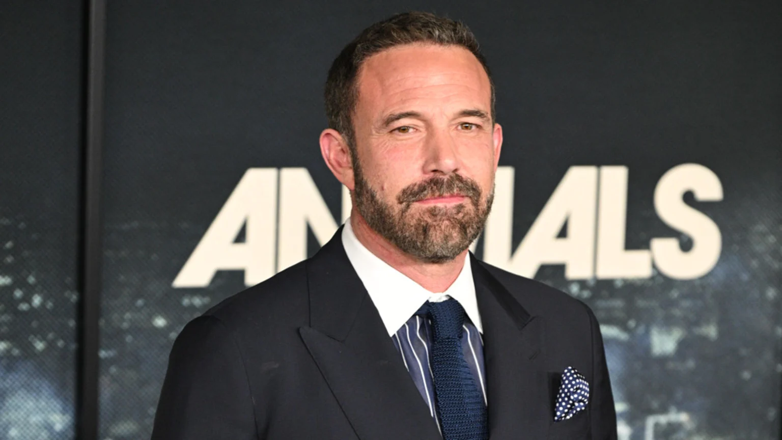 Ben Affleck, with a beard and short dark hair, wears a dark suit, white shirt, blue striped tie, and a polka dot pocket square, standing against a dark background with large white letters that partially spell "ANIMALS."