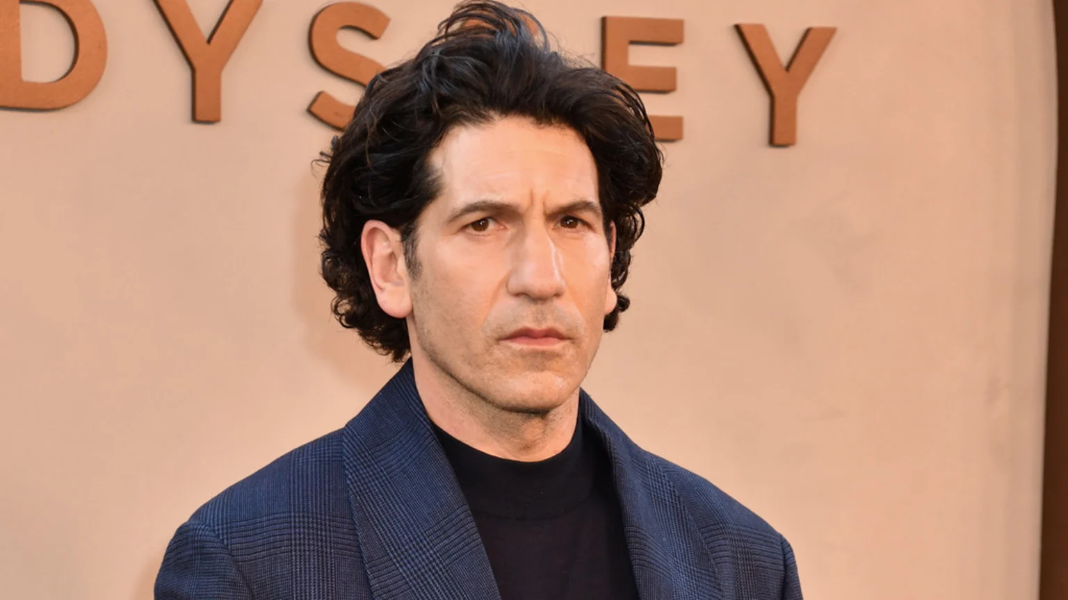 Jon Bernthal, with dark, curly hair and a serious expression, wears a dark blue patterned blazer over a black turtleneck, looking directly at the camera, with part of an "ODYSSEY" sign in the background.