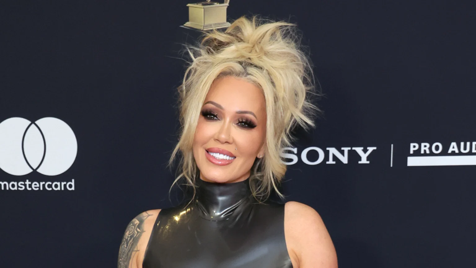 A blonde woman with a messy high bun and heavy makeup smiles at the camera wearing a shiny black sleeveless top. The indoor portrait shows a detailed tattoo on her left arm against a dark backdrop featuring Mastercard and Sony logos and a small golden trophy-like object above her head.