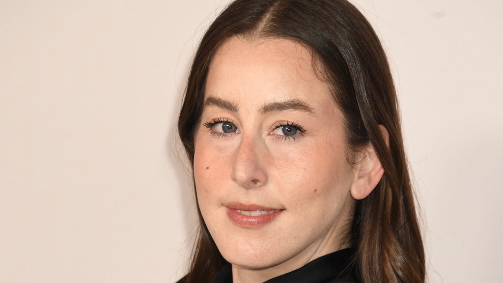 A woman with long dark hair, blue eyes, and a faint smile looks directly at the viewer in a close-up portrait against a light background.