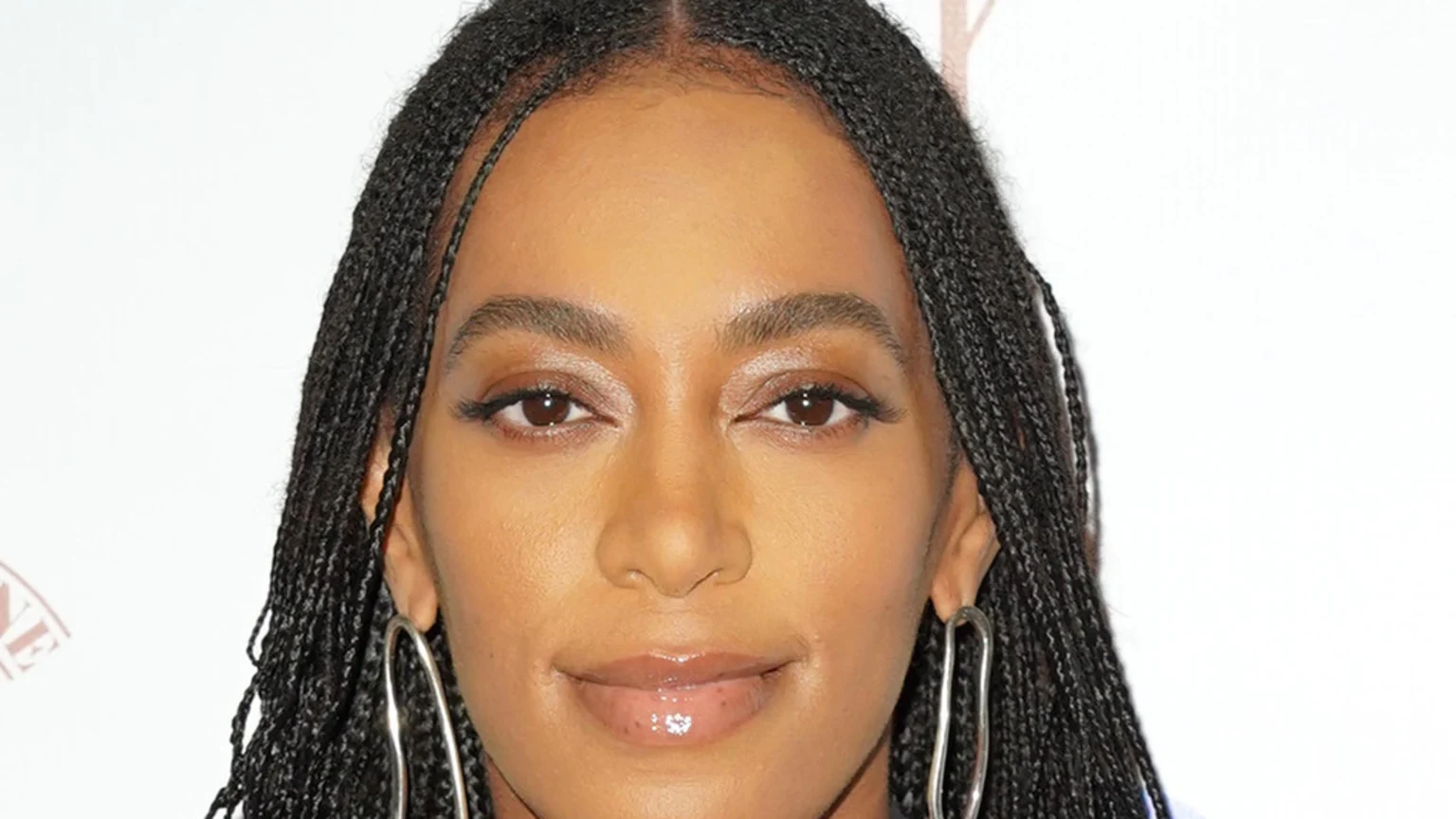 Close-up portrait of Solange Knowles showing her braided hair parted down the middle, defined eyebrows, winged eyeliner, glossy lips, and large silver hoop earrings.