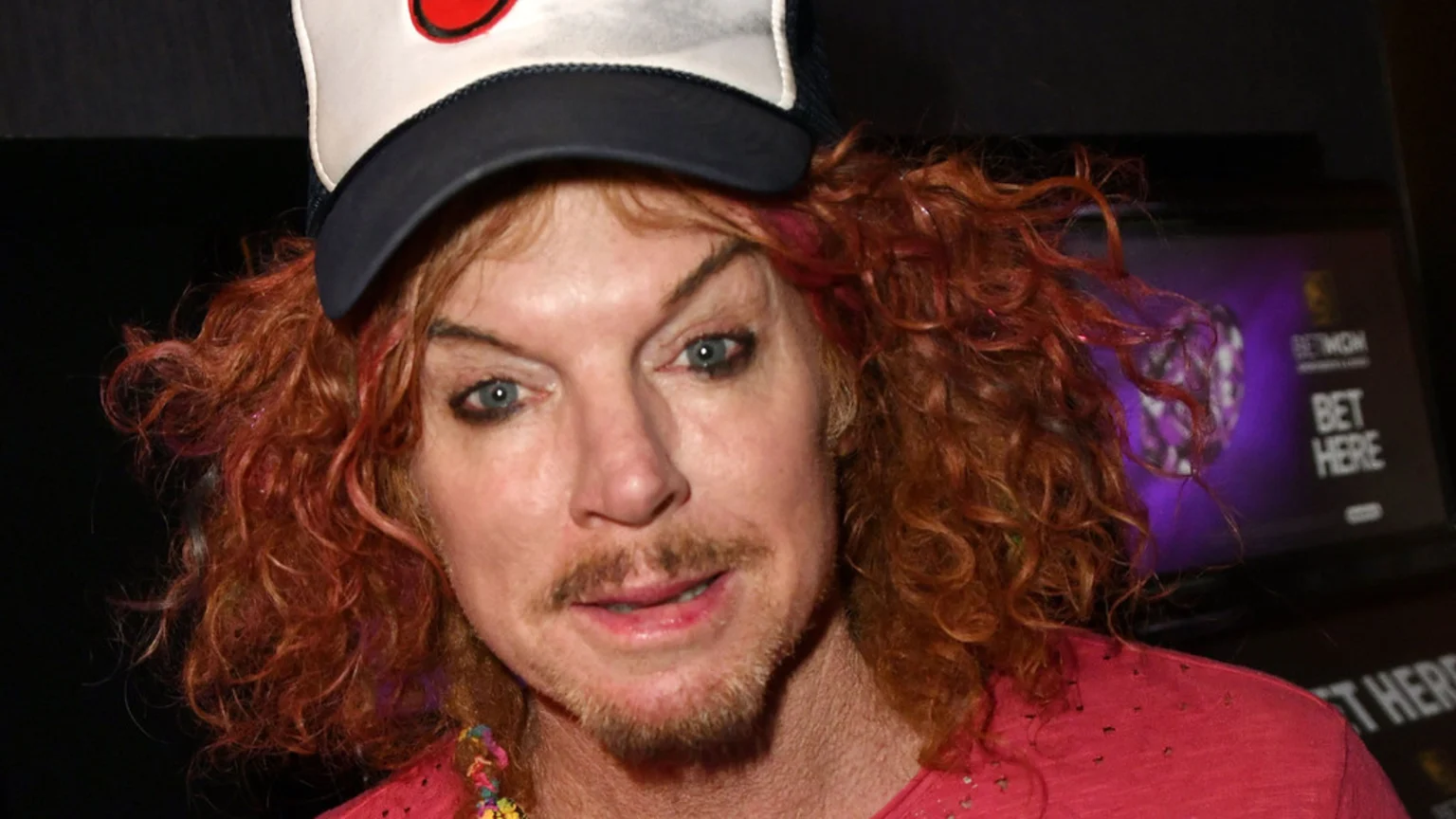 Close-up of comedian Carrot Top, with curly red hair, wearing a white baseball cap, pink shirt, and a colorful necklace, looking forward. A blurry "BETMGM" sign is in the background.