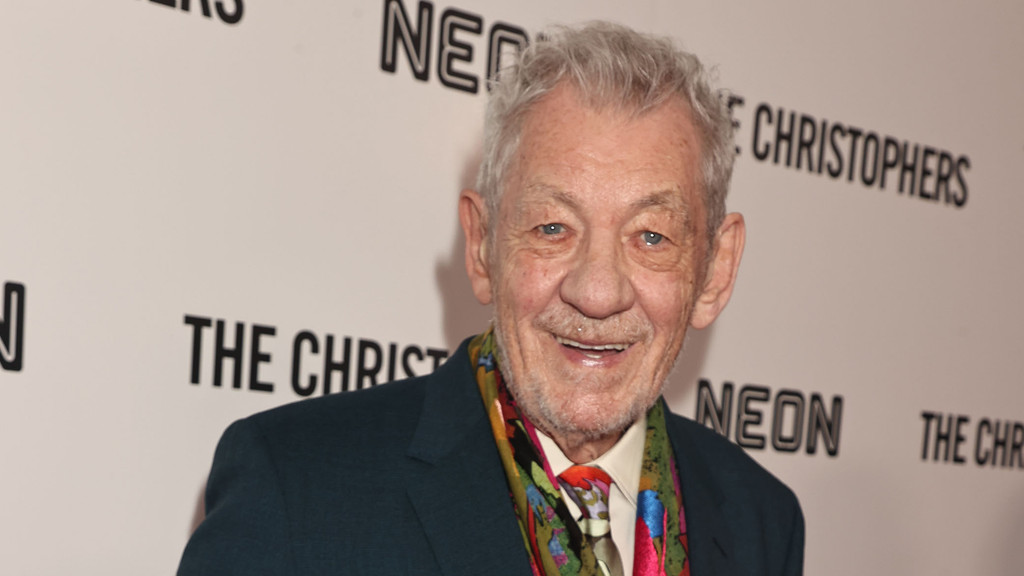 A smiling elderly man with grey hair and blue eyes, wearing a dark suit, colorful tie, and scarf, stands in front of a white wall with black text.
