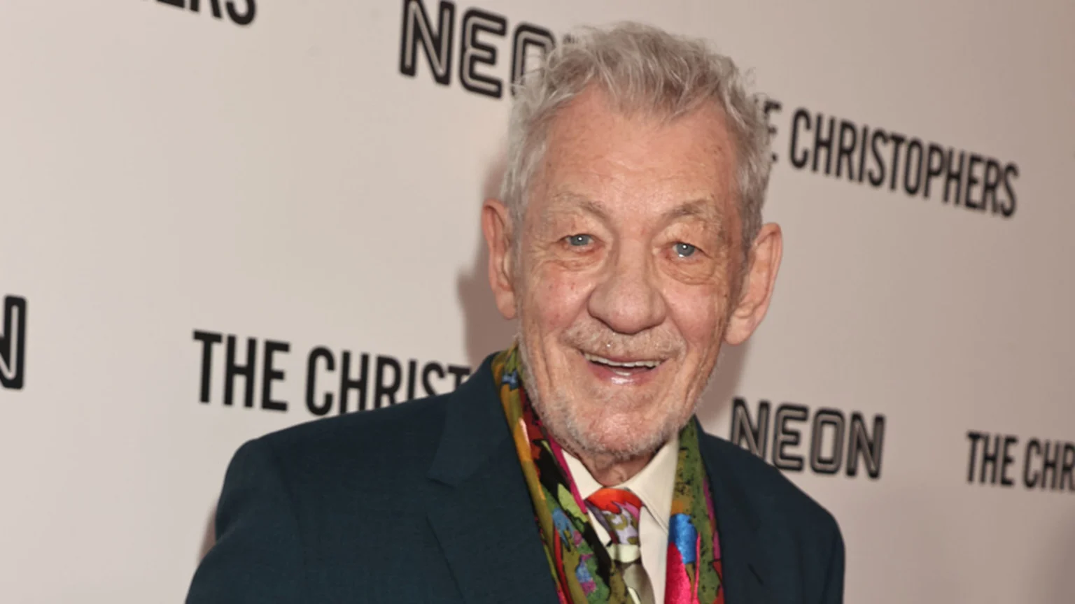 A smiling elderly man with grey hair and blue eyes, wearing a dark suit, colorful tie, and scarf, stands in front of a white wall with black text.