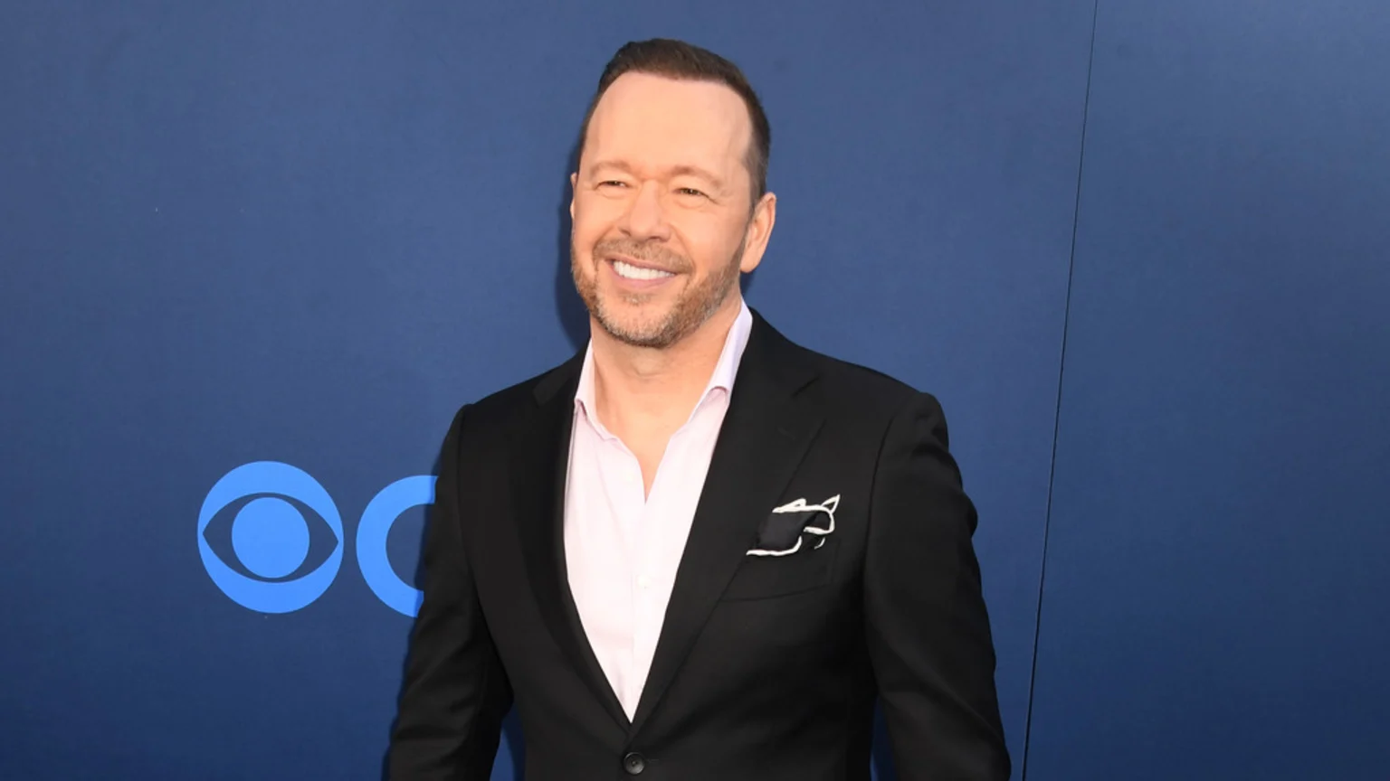 Donnie Wahlberg smiles, wearing a black suit jacket, a light pink shirt, and a patterned pocket square, against a blue background with parts of the CBS logo.