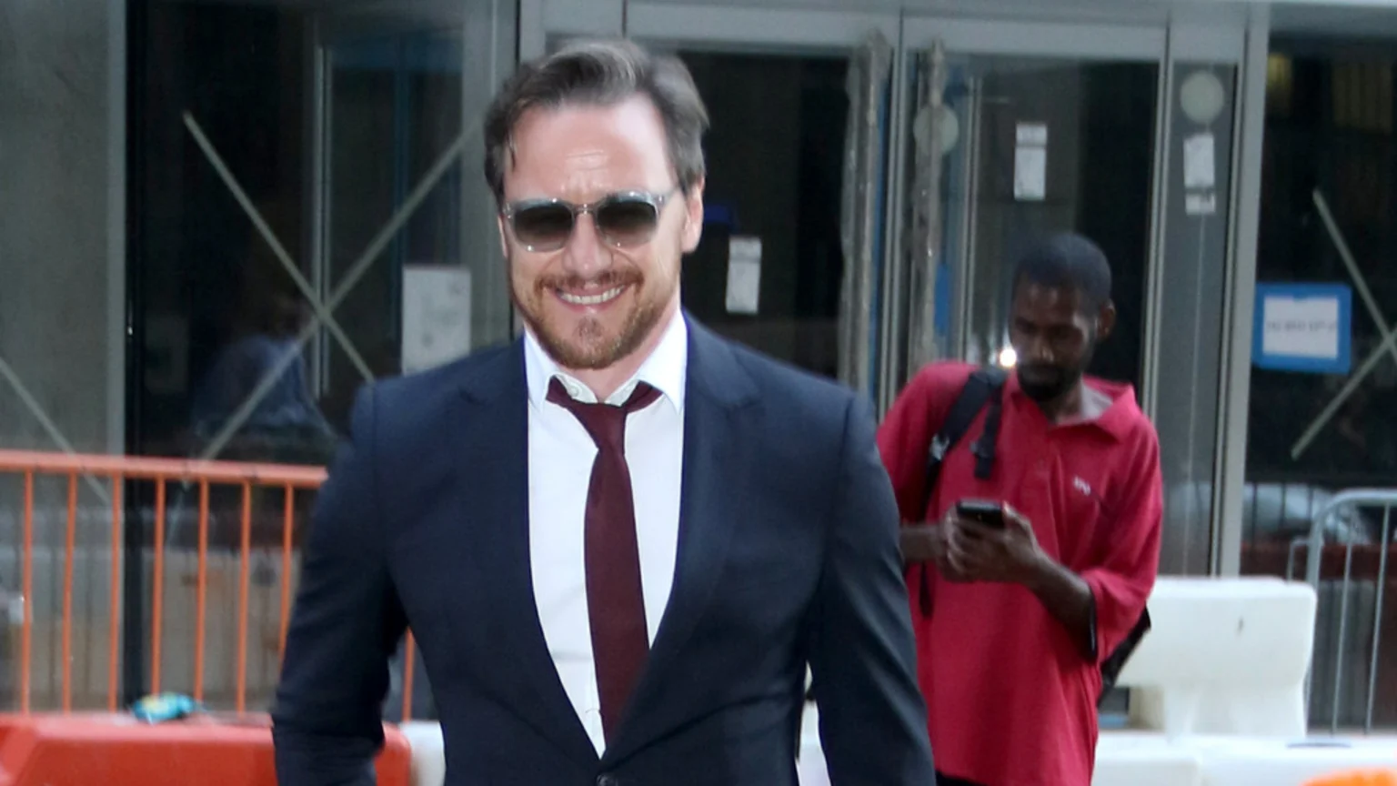 James McAvoy smiles while wearing sunglasses, a dark suit, white shirt, and maroon tie, walking in front of a building with another man in a red shirt visible behind him.