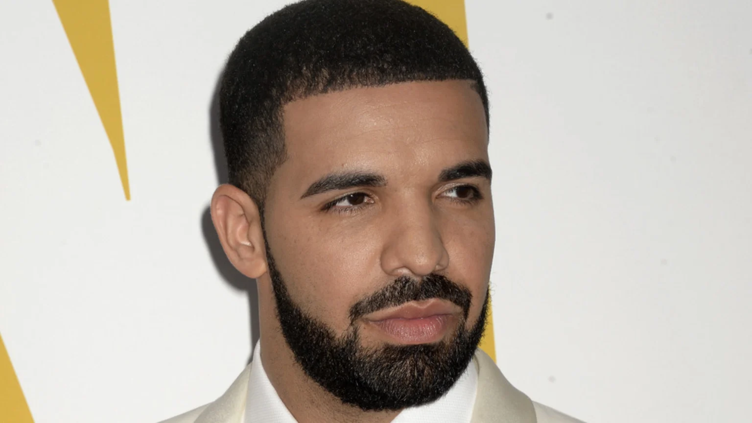 Rapper Drake looks to his right, wearing a light suit and white shirt, with a short haircut and full beard, against a white and yellow background.