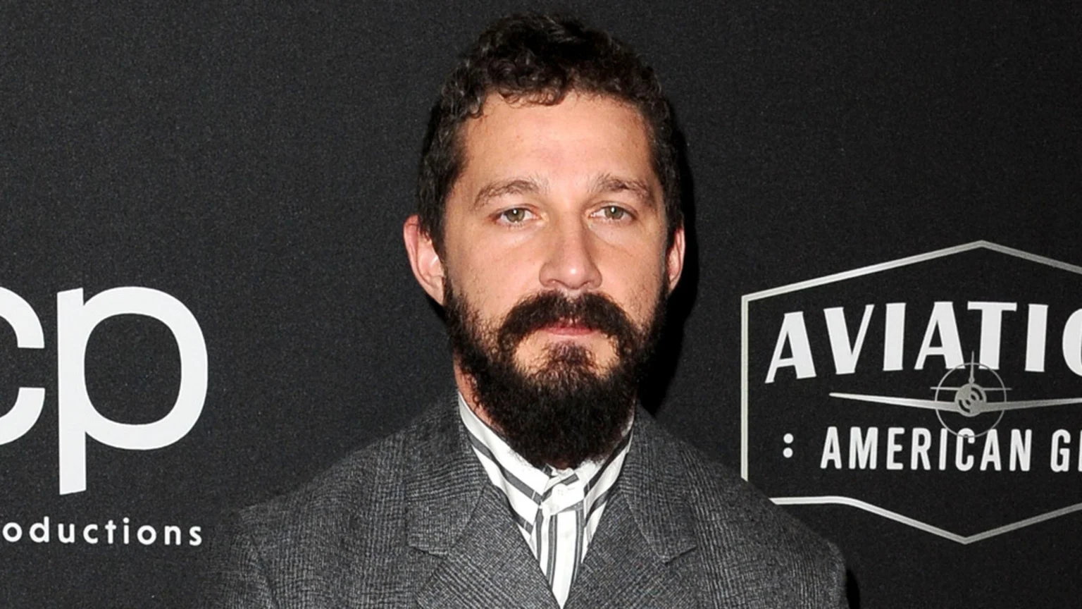 Actor Shia LaBeouf with a dark beard and curly hair, wearing a grey jacket and striped shirt, looks at the camera against a dark background with logos.