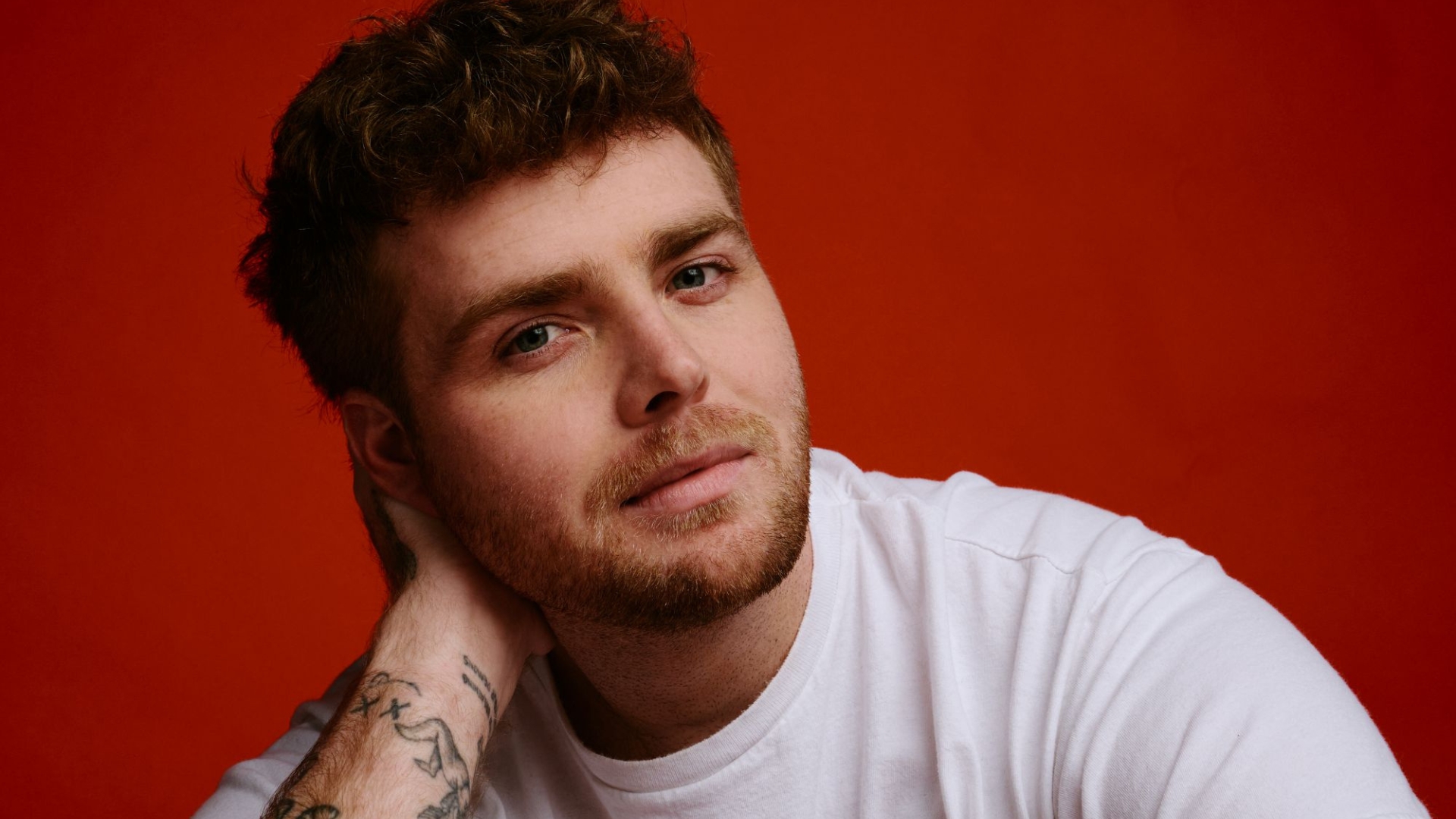 Close-up of a man with reddish-brown curly hair, green eyes, and beard, wearing a white t-shirt, posing with his tattooed arm behind his head against a red background.